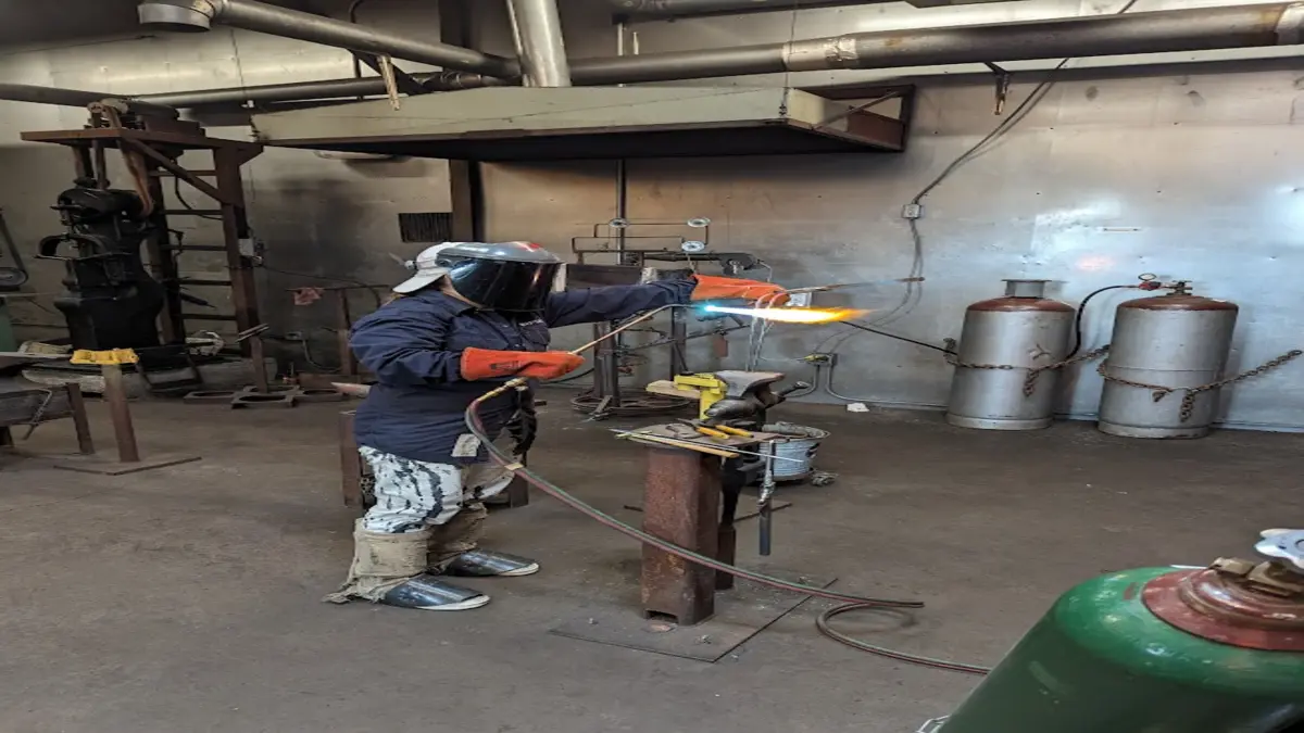 Person in protective gear heating a metal bar with a torch in an industrial workshop.